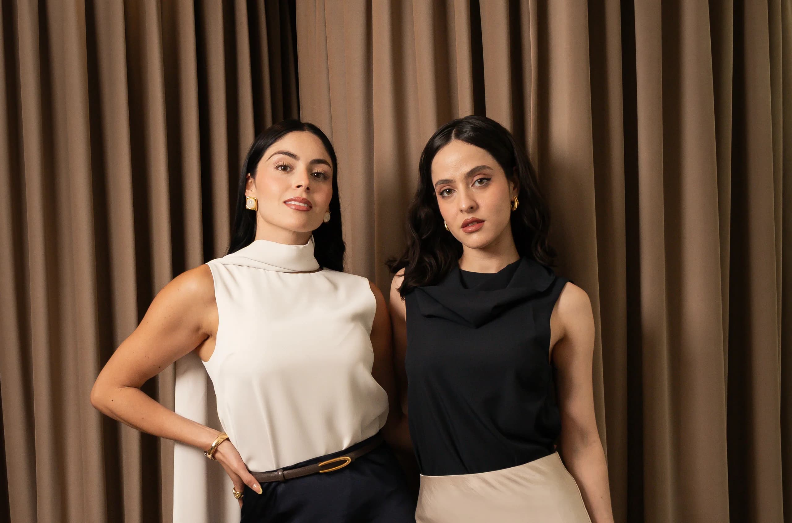 Dos mujeres frente a una cortina color arena: una con top blanco de cuello alto y falda azul marino, la otra con top negro drapeado y falda satinada beige.